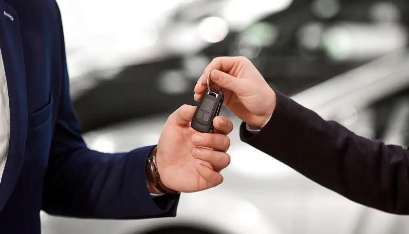 Deux personnes échangent une clé de voiture noire en gros plan, avec une voiture floue en arrière-plan.