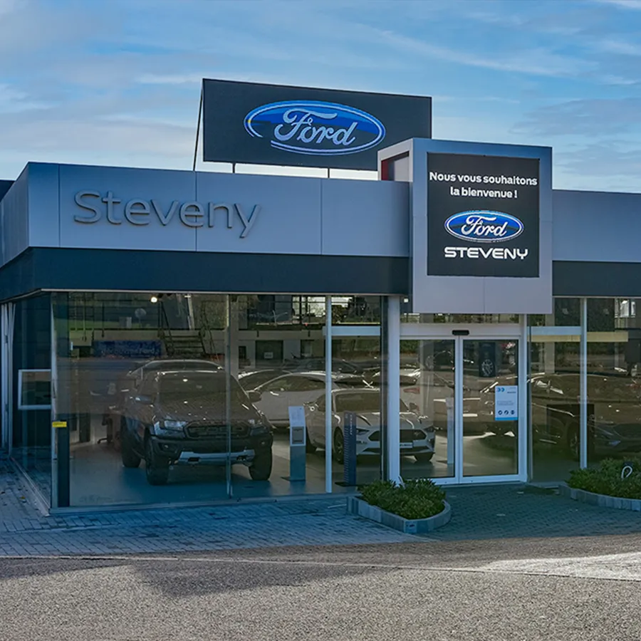 Façade de la concession Ford Steveny à Namur avec des voitures exposées derrière des vitrines et un ciel bleu.