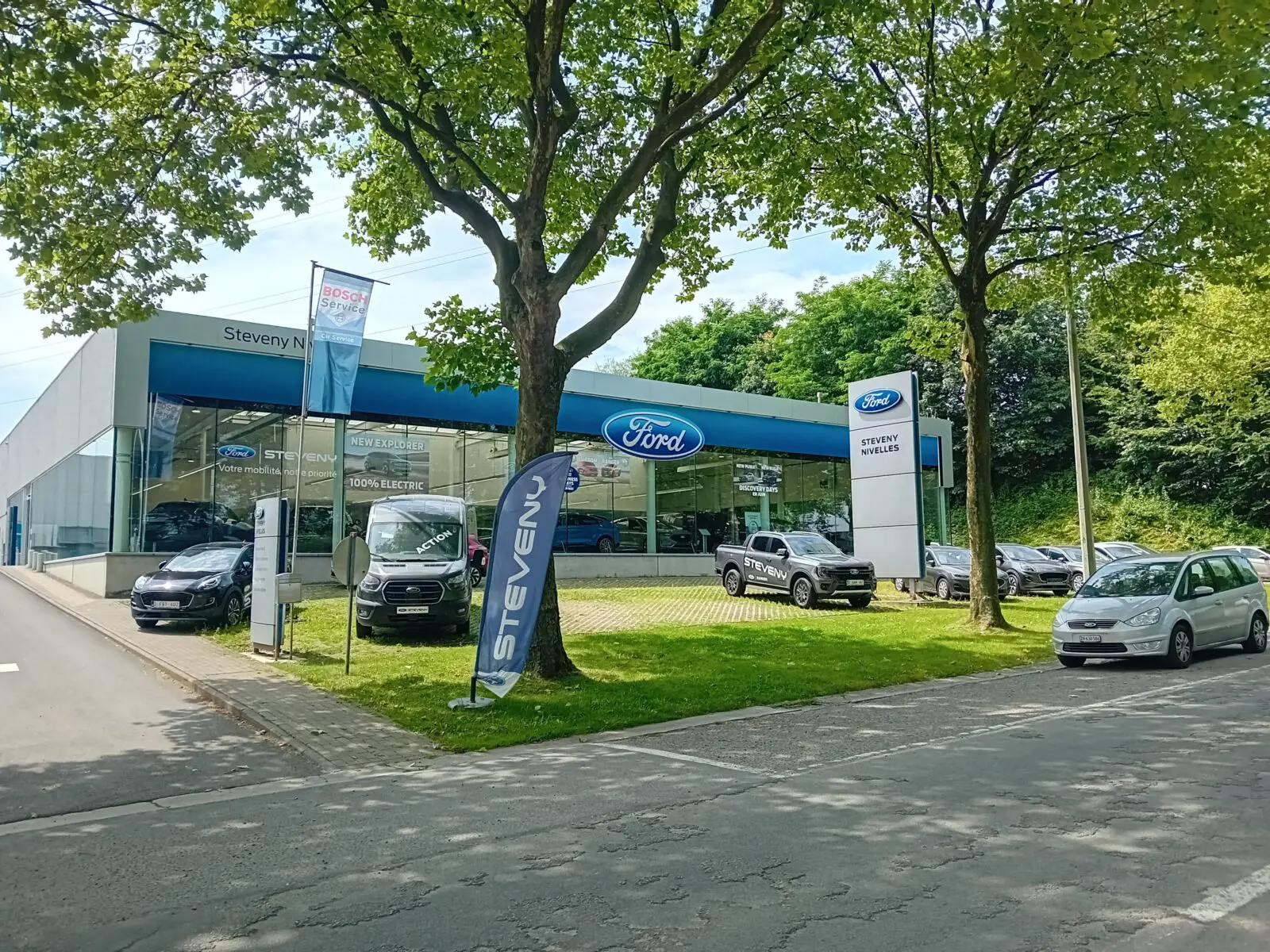 Ford_Steveny_Nivelles-1 Concession Ford Steveny à Nivelles avec plusieurs voitures stationnées à l'extérieur, entourée d'arbres et de bannières publicitaires.