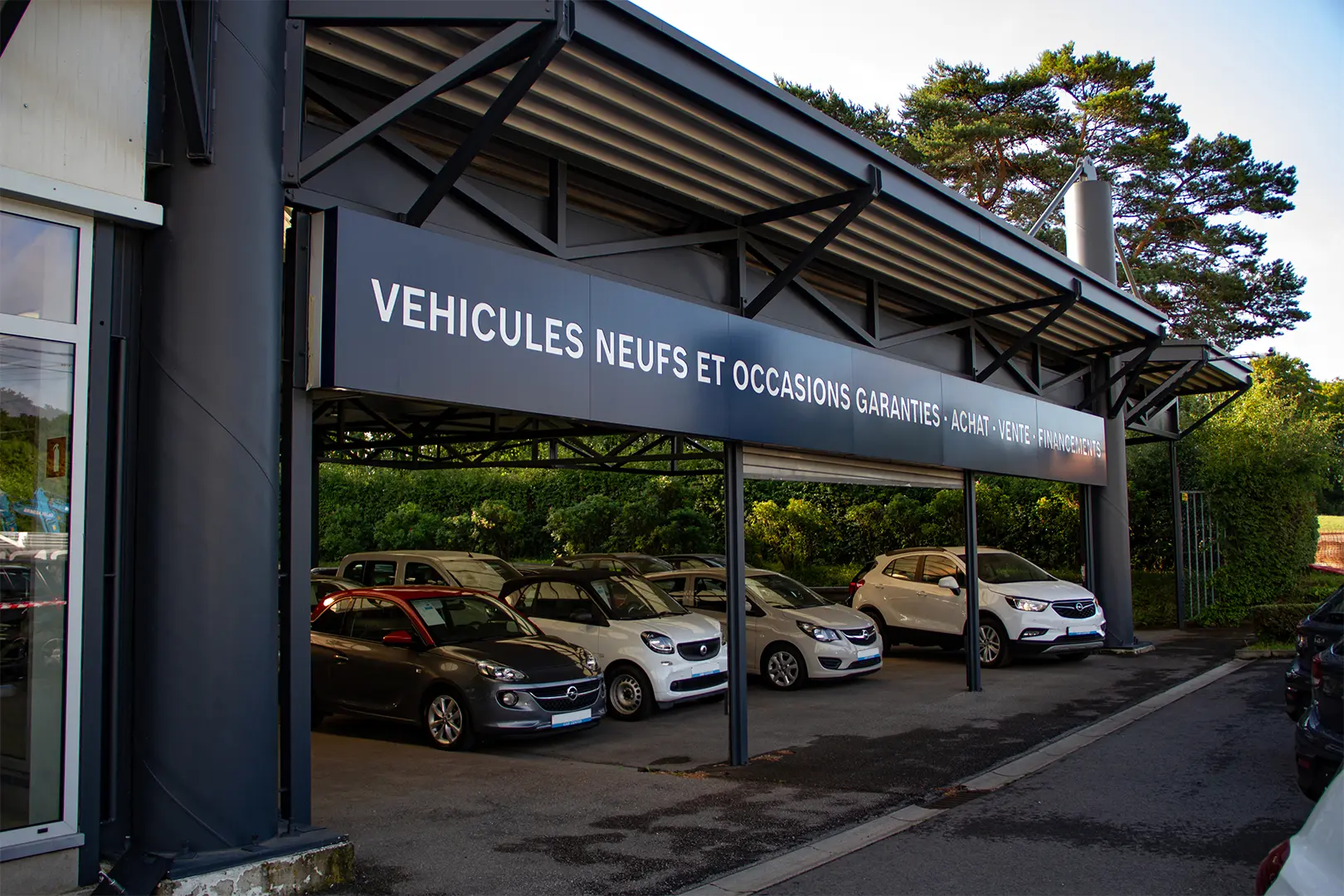 Un abri de voitures avec des véhicules Opel gris et blancs, sous un panneau annonçant des véhicules neufs et d'occasion garantis.