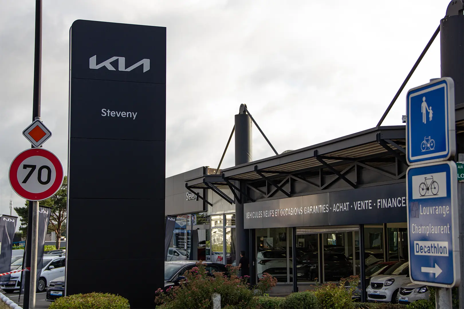 Panneau Kia noir et bâtiment de concession Steveny avec voitures visibles à l'intérieur, panneaux de signalisation à proximité.