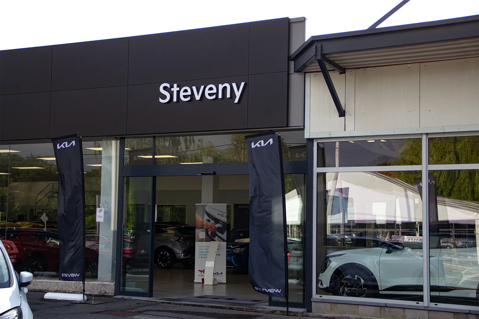 Façade de la concession Steveny avec des drapeaux Kia et une voiture blanche visible à travers la vitre.