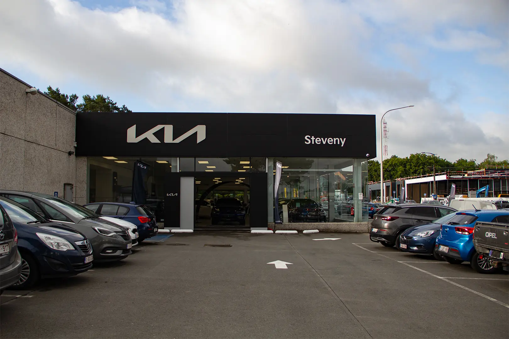 Façade de la concession Steveny avec logo Kia, voitures garées à l'extérieur sous un ciel nuageux.