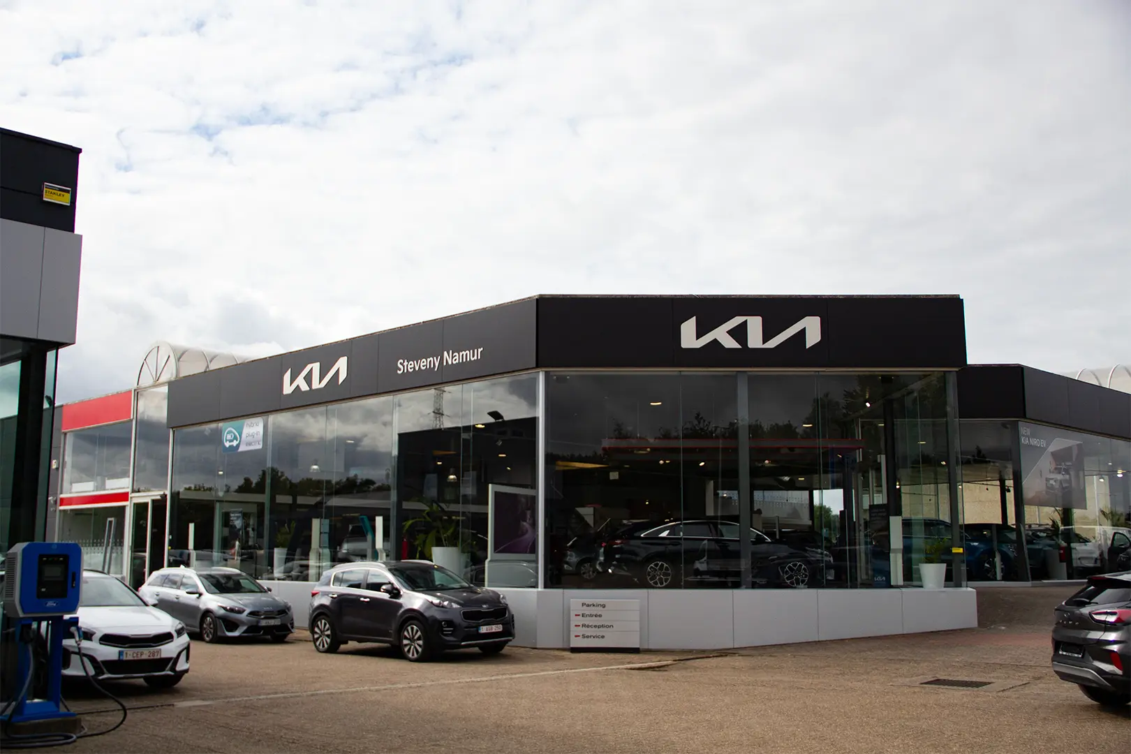 Concession Kia Steveny Namur avec plusieurs voitures stationnées devant, bâtiment moderne en verre sous un ciel nuageux.