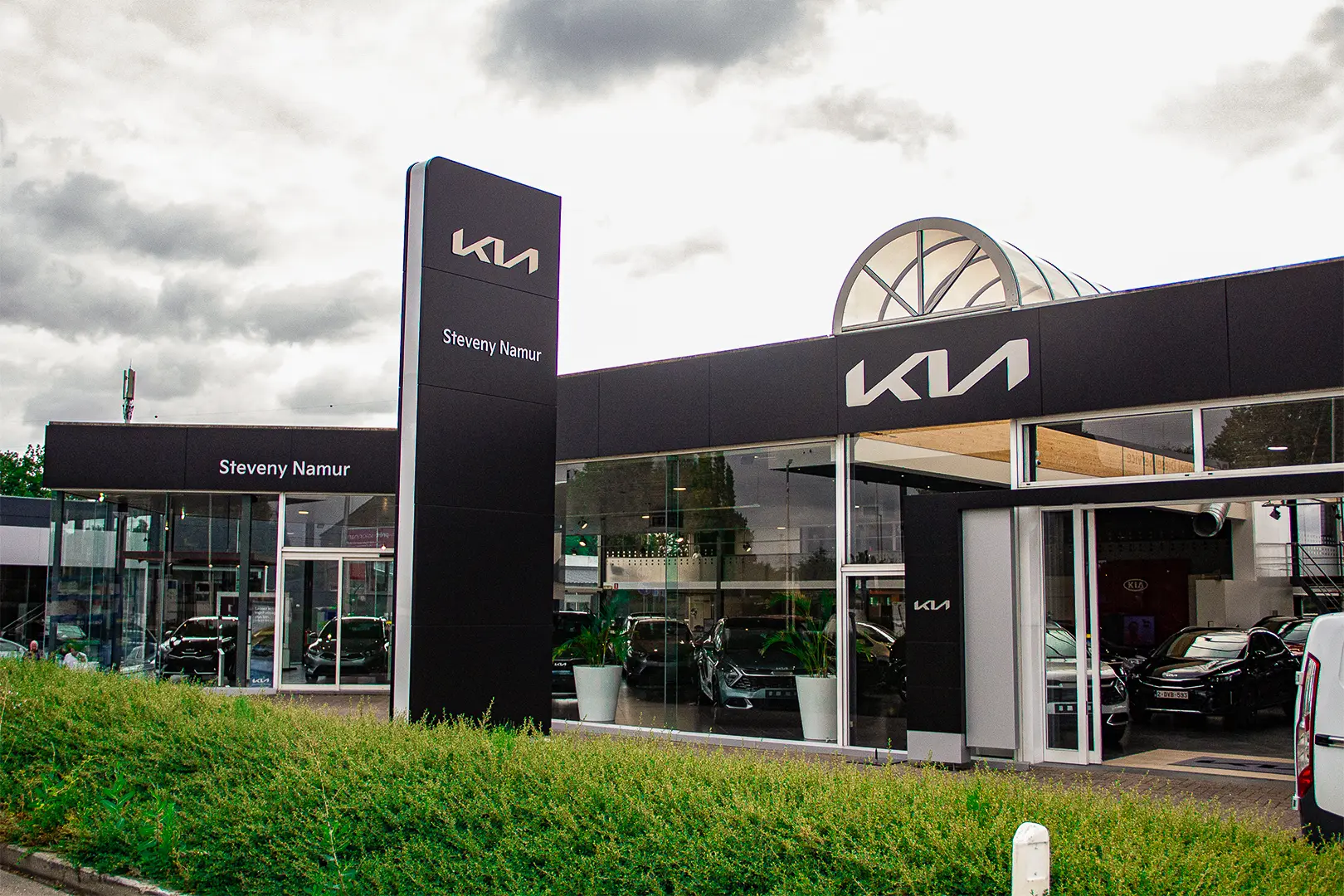 Façade de la concession Kia Steveny Namur avec des voitures exposées à l'intérieur, sous un ciel nuageux.