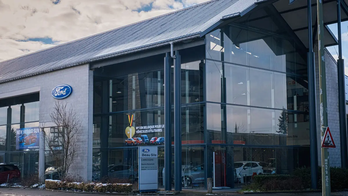Concession Ford à Arlon avec façade vitrée, logo Ford bleu et affiches promotionnelles visibles à l'extérieur.