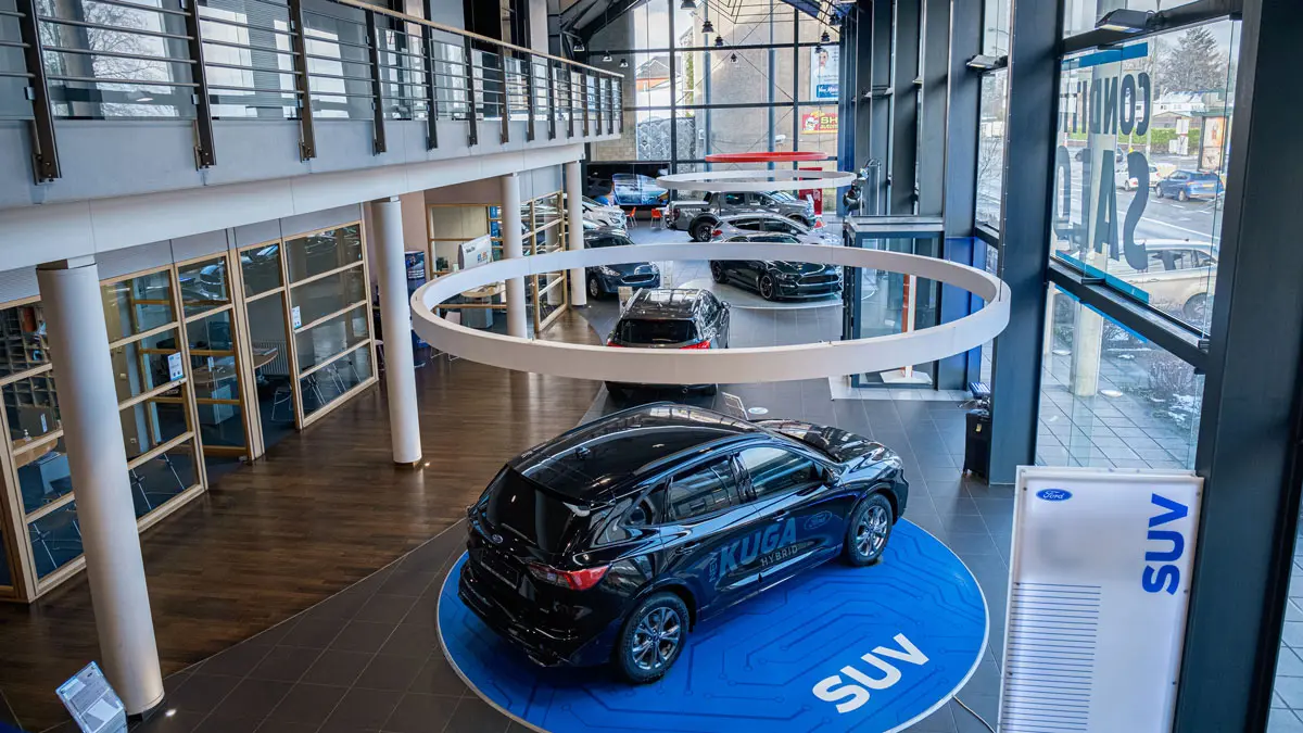 Salon d'exposition Ford avec plusieurs véhicules, dont un Ford Kuga noir sur un podium bleu marqué "SUV", intérieur moderne et spacieux.