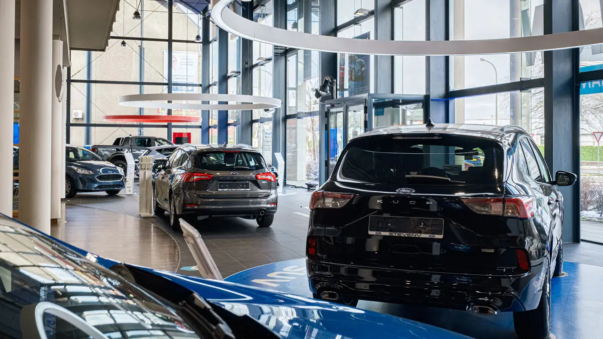 Des voitures Ford, dont un Kuga noir et un Focus gris, sont exposées dans une concession lumineuse avec de grandes fenêtres.