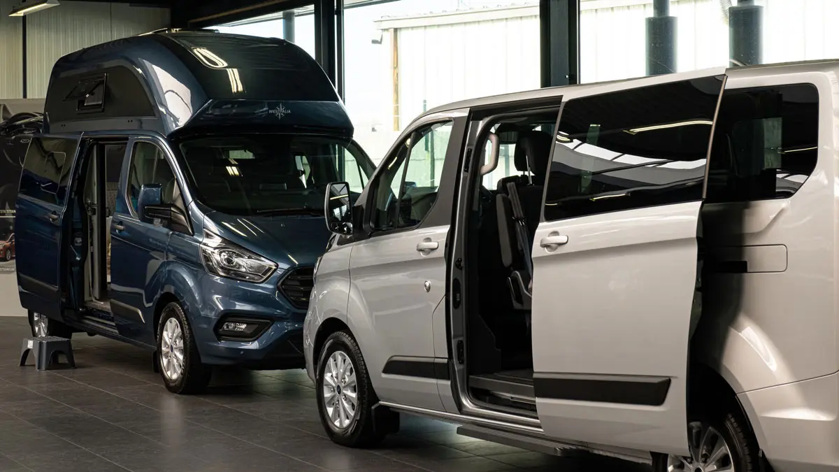 Deux fourgons Ford, l'un bleu et l'autre blanc, sont exposés dans une concession automobile, portes ouvertes.
