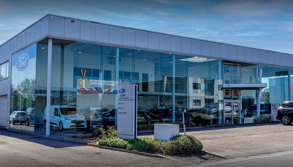 Concession Ford Steveny à Braine-l'Alleud avec façade vitrée, logo Ford et véhicules visibles à l'intérieur.
