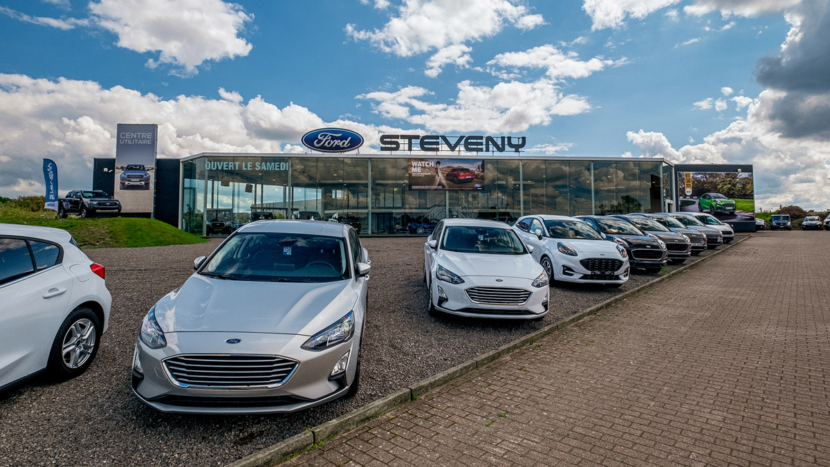 Concession Ford de Charleroi Airport Concession Ford Steveny à Charleroi avec plusieurs voitures Ford argentées alignées devant le bâtiment vitré sous un ciel partiellement nuageux.
