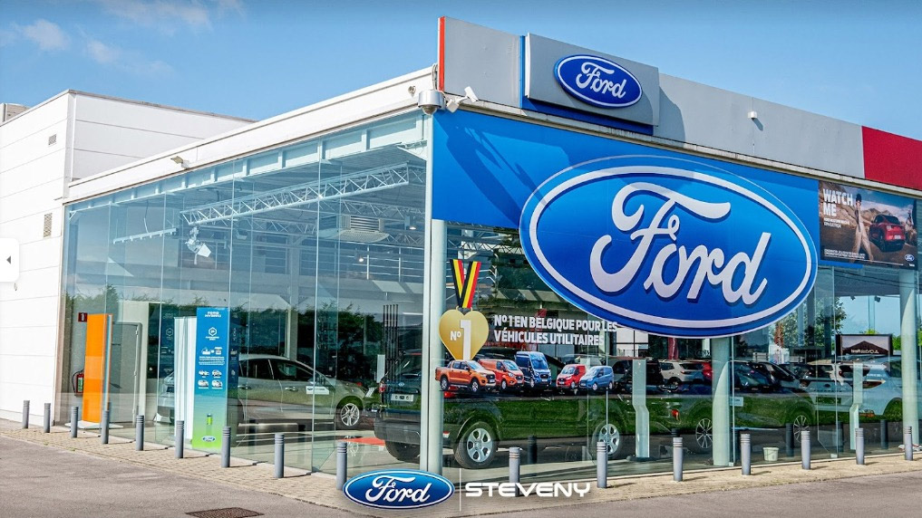 Concession Ford à Dinant avec une façade vitrée, grand logo Ford bleu et voitures exposées à l'intérieur.