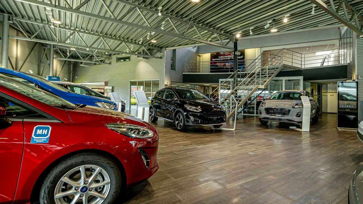 Intérieur d'une concession Ford à Gembloux avec des voitures rouges, bleues, noires et grises exposées sous un plafond métallique.