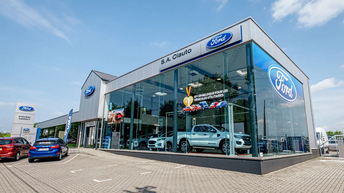 concession-ford-hannut-1 Concession Steveny Ford à Hannut avec plusieurs véhicules exposés derrière une façade vitrée. Panneau Ford visible à l'extérieur.