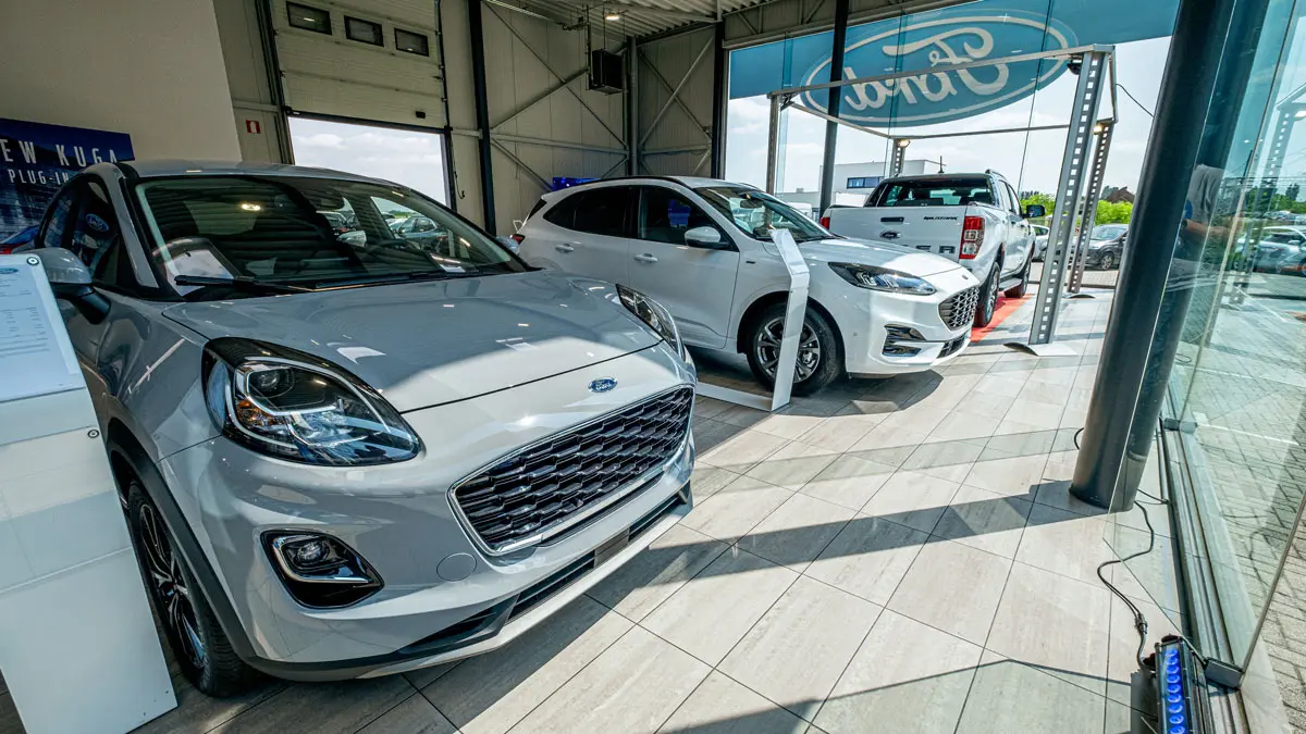 concession-ford-hannut-2 Trois voitures Ford grises et blanches sont exposées dans une concession avec un logo Ford visible à l'extérieur.