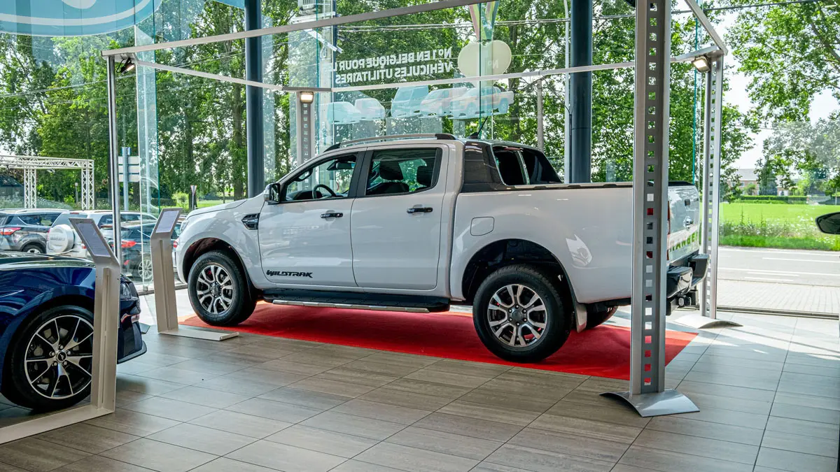 concession-ford-hannut-3 Un pick-up Ford blanc est exposé sur un tapis rouge dans une concession automobile avec de grandes fenêtres.