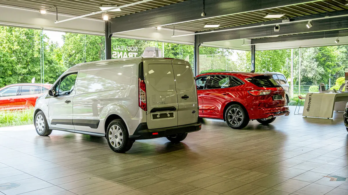 concession-ford-hannut-4 Un utilitaire Ford blanc et un SUV Ford rouge sont exposés dans une concession automobile lumineuse avec vue sur l'extérieur verdoyant.
