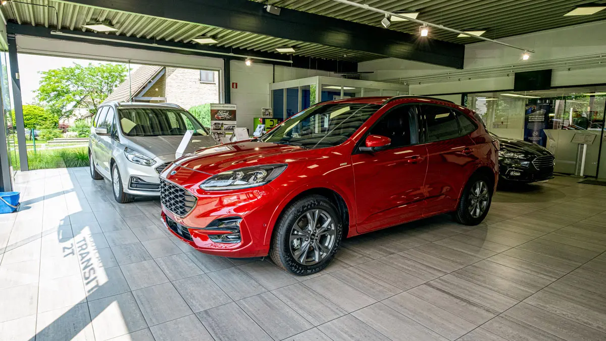 concession-ford-hannut-5 Deux voitures Ford, une rouge et une grise, sont exposées dans une concession automobile moderne avec de grandes fenêtres.