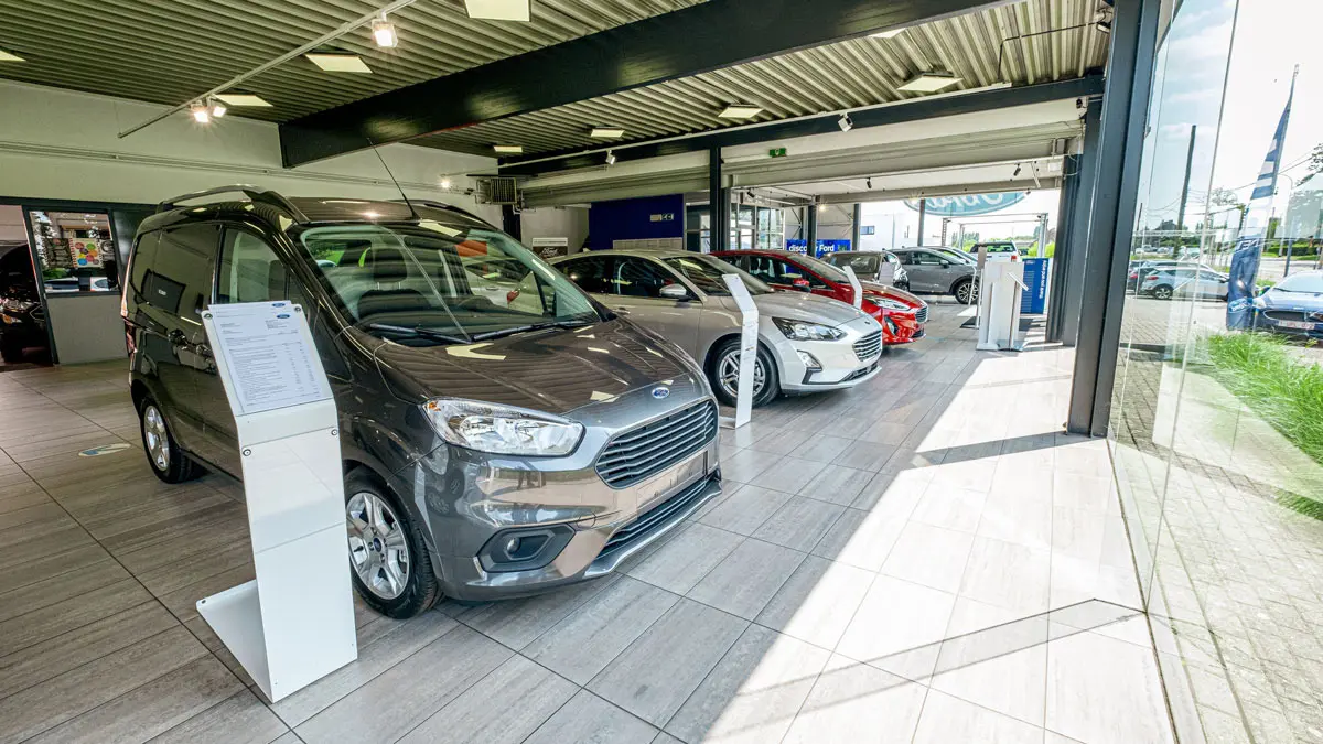 concession-ford-hannut-6 Une Ford gris foncé est exposée à l'intérieur d'une concession, entourée d'autres modèles de voitures alignés.