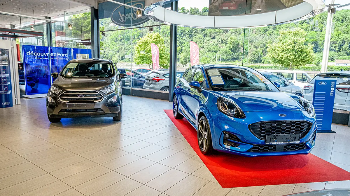 Deux voitures Ford, un modèle gris et un modèle bleu, sont exposées dans une concession avec des vitrines et un tapis rouge.