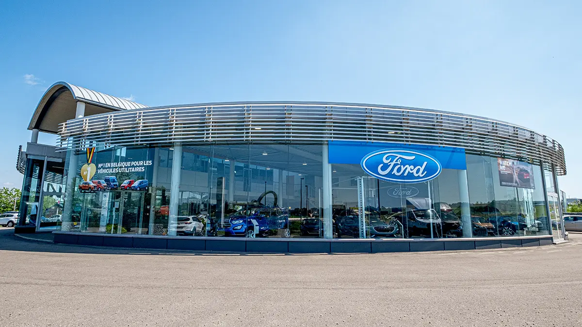 concession-ford-marche-1 Concession Ford avec façade vitrée, affichant des véhicules exposés à l'intérieur sous un ciel bleu.