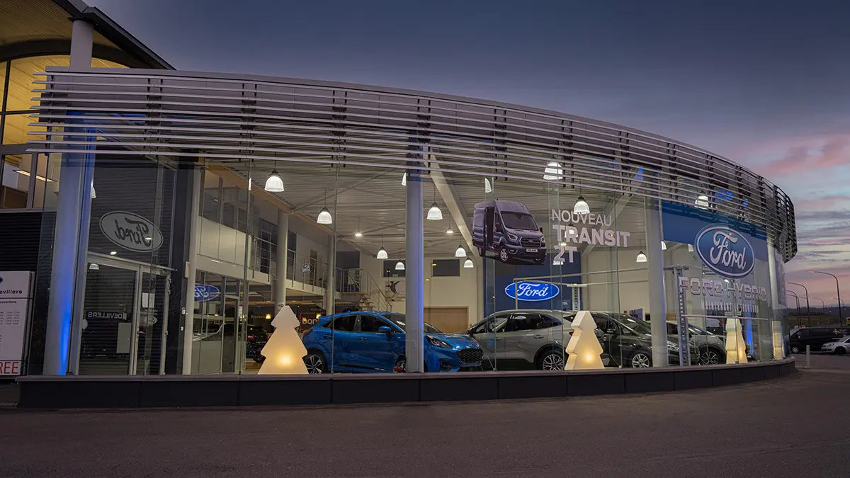 concession-ford-marche-2 Concession Ford avec voitures exposées, dont un modèle bleu, éclairée la nuit avec des décorations lumineuses en forme de sapins.