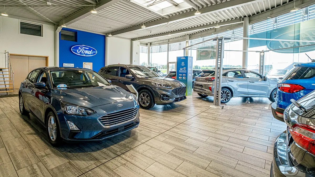 concession-ford-marche-5 Voitures Ford exposées dans une concession avec un logo Ford bleu sur le mur, éclairage naturel et sol carrelé.
