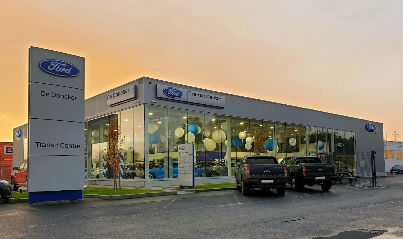 concession-ford-mons-1 Concession Ford à Mons avec enseigne "Transit Centre" et plusieurs véhicules stationnés devant, ciel au crépuscule en arrière-plan.