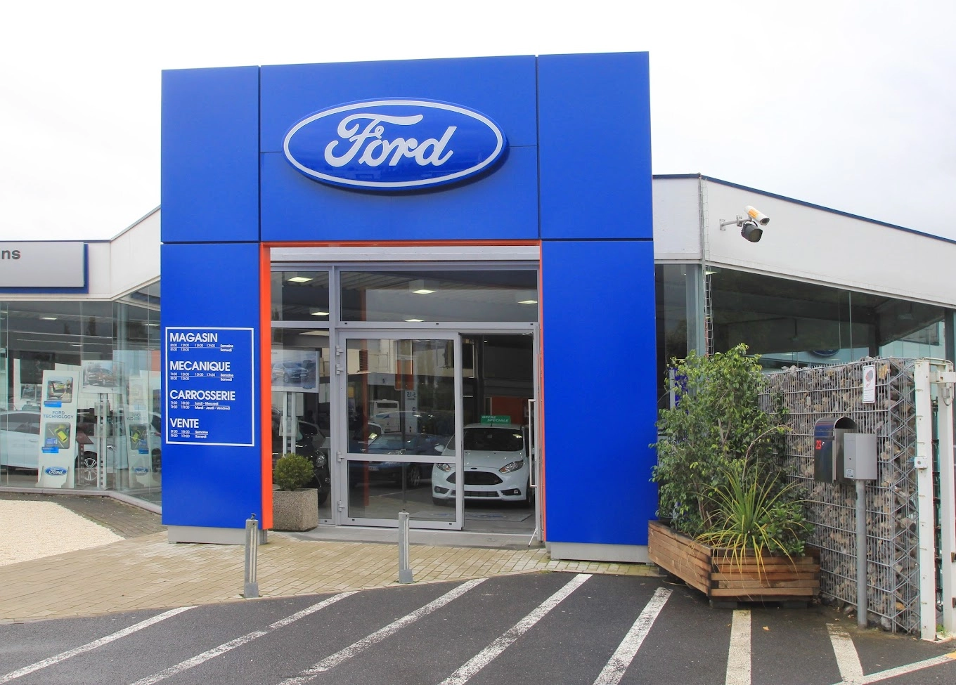 concession-ford-mons-3 Entrée de la concession Ford à Mons avec façade bleue et enseigne Ford, voiture blanche visible à l'intérieur.