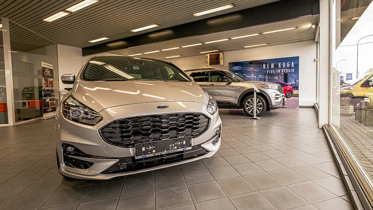 concession-ford-nivelles-3 Voitures Ford exposées dans une concession à Nivelles, avec un modèle argenté au premier plan et un panneau publicitaire en arrière-plan.