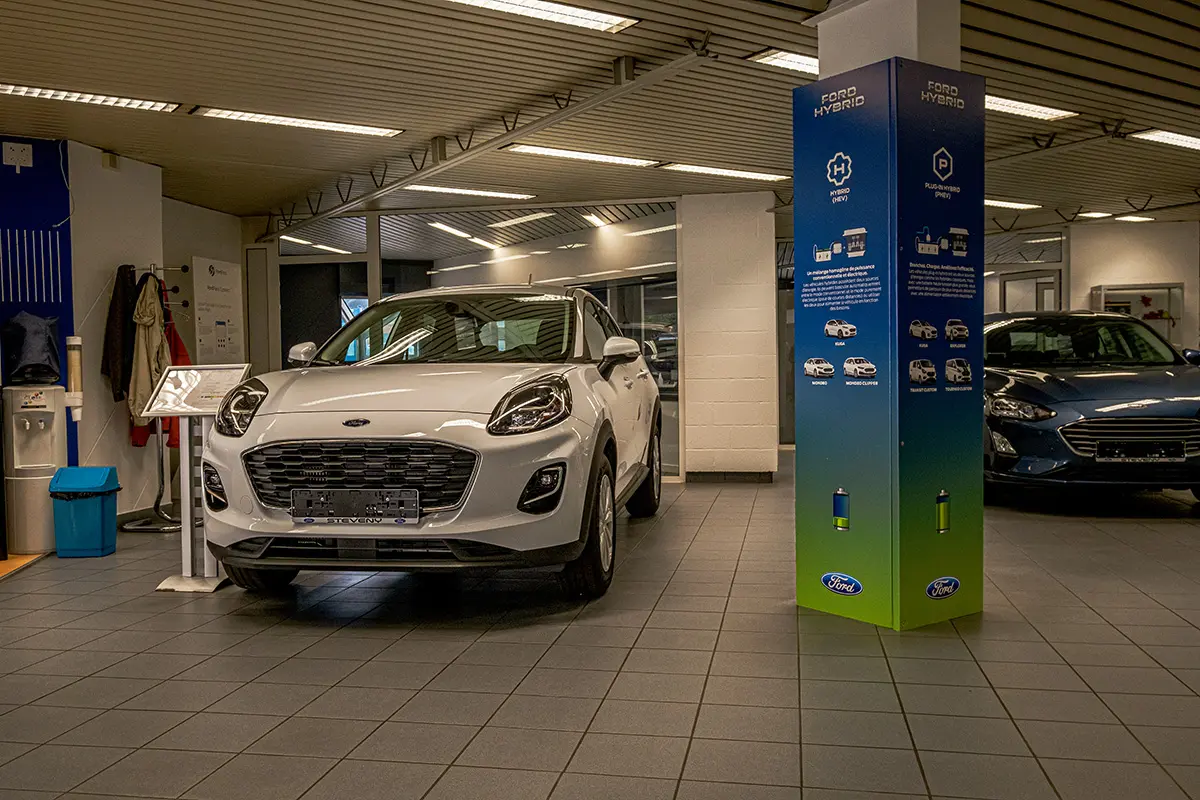 concession-ford-nivelles-4 Voiture Ford blanche exposée dans une concession Steveny, avec un panneau d'information sur les modèles hybrides à droite.