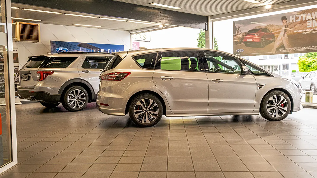 concession-ford-nivelles-6 Deux voitures Ford argentées stationnées dans une concession automobile avec un sol carrelé gris et des affiches publicitaires.