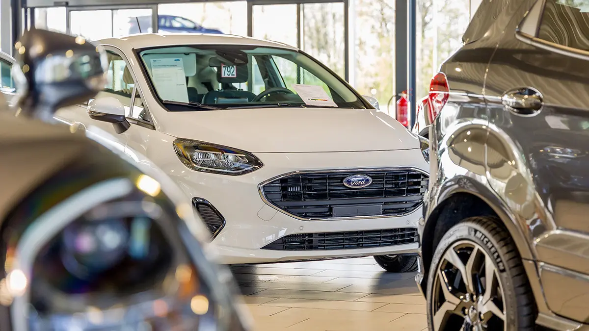 concession-ford-somzee-6 Une Ford blanche est exposée dans une concession, entourée d'autres véhicules, sur un sol carrelé clair.