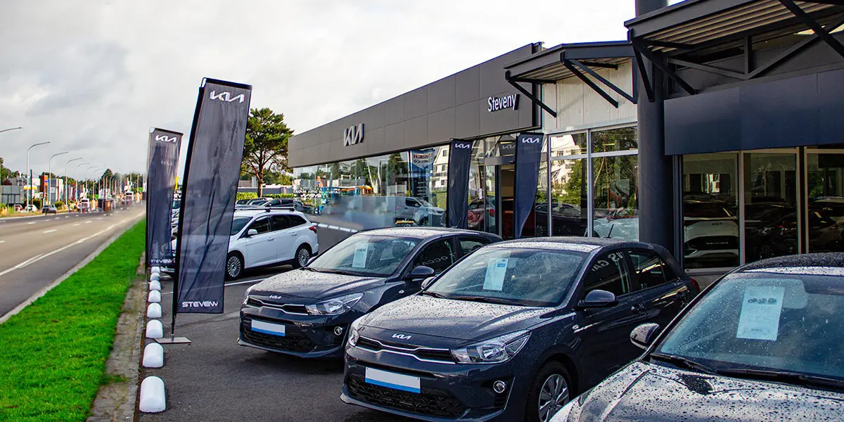 Concession Steveny avec des voitures Kia noires garées devant, des drapeaux Kia visibles, près d'une route.