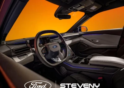 Intérieur d'une voiture Ford avec volant et tableau de bord modernes, fond orange vif, logo Steveny visible.