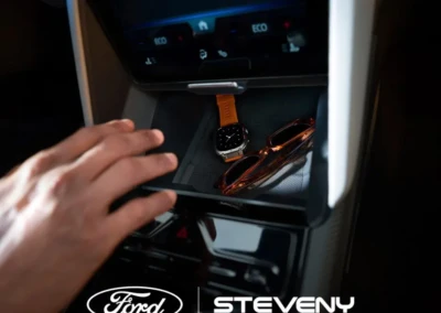 Main posée sur le tableau de bord d'une voiture Ford, avec une montre orange et des lunettes de soleil dans le rangement.