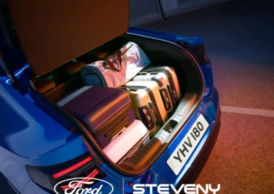 Coffre d'une voiture Ford bleu rempli de valises et d'un sac, avec logo Steveny visible.