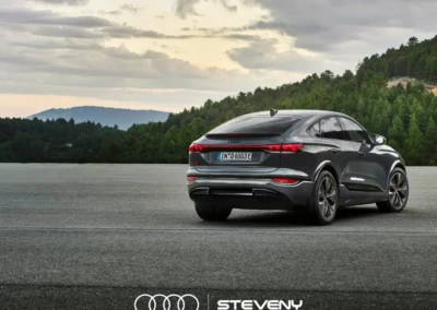 Audi Q6 Sportback e-tron gris sur une route, entourée de forêt et ciel nuageux, avec logo Steveny en bas.