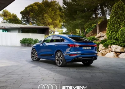 Audi Q6 Sportback e-tron bleu stationné sur une allée pavée, entourée de verdure et d'arbres, avec logo Steveny en bas.