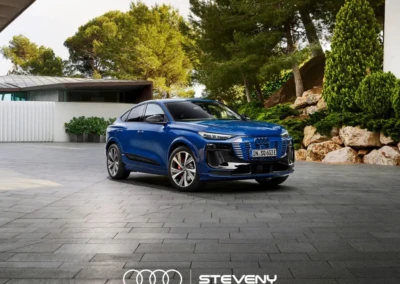 Audi Q6 Sportback e-tron bleu garée sur une allée pavée, entourée de végétation et d'un bâtiment moderne.