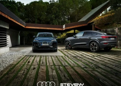 Deux Audi Q6 Sportback e-tron noires stationnées sur une terrasse en bois, entourées de végétation et d'une structure moderne.