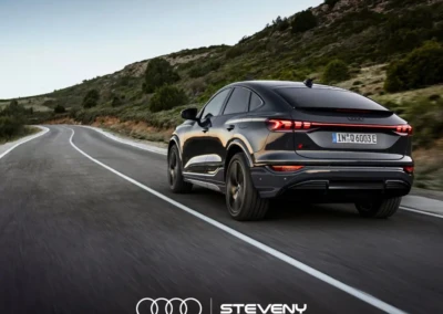 Audi Q6 Sportback e-tron gris foncé roulant sur une route sinueuse bordée de végétation, logo Steveny en bas.