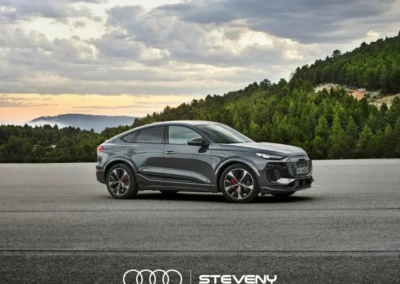 Audi Q6 Sportback e-tron gris stationné sur une route devant une forêt, sous un ciel nuageux. Logo Steveny visible.