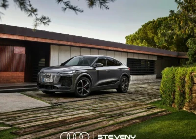 Voiture Audi Q6 Sportback e-tron grise stationnée devant une maison moderne, entourée de verdure et d'arbres.