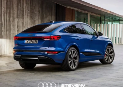 Voiture bleue Audi Q6 Sportback e-tron garée devant un bâtiment moderne en béton, vue arrière trois-quarts.