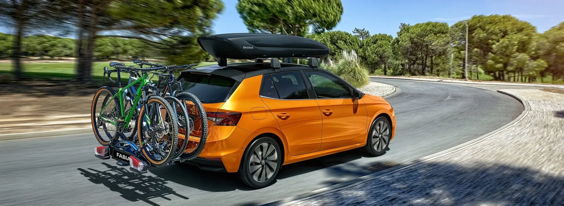 Voiture orange Skoda avec un coffre de toit noir et trois vélos accrochés à l'arrière, roulant sur une route bordée d'arbres.
