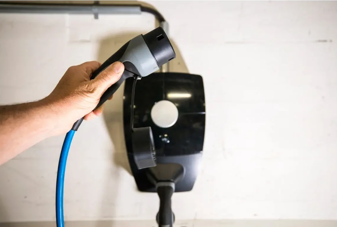 Une main tient une prise de charge électrique noire et bleue devant une borne murale noire.