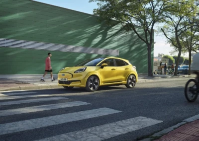 Voiture Ford Puma 2024 jaune stationnée près d'un passage piéton, avec un mur vert en arrière-plan et des arbres.