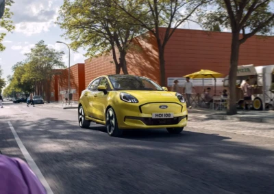 Voiture Ford Puma 2024 jaune roulant sur une route bordée d'arbres et de bâtiments rouges par une journée ensoleillée.