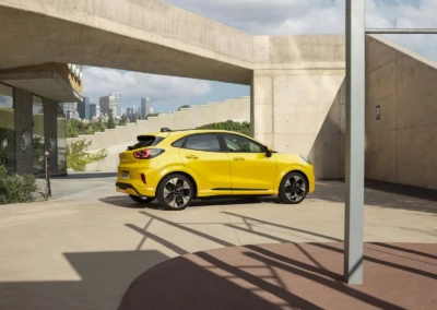 Voiture Ford Puma Gen-E 2024 jaune garée dans un espace urbain avec des bâtiments en arrière-plan.